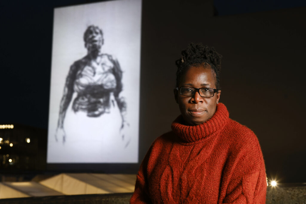 Rosamund Adoo-Kissi-Debrah in front of a projection of Breathe For Ella, a new iteration of Dryden Goodwin's artwork depicting Rosamund, the mother of Ella Adoo-Kissi-Debrah, fighting for breath at the Rambert Building, in London. Credit: David Parry/PA Images via Getty Images
