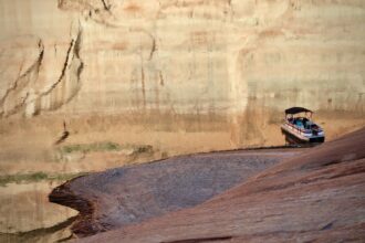 A pontoon boat is tied up at the shore of a recently-revealed beach in one of Lake Powell's side canyons on April 10, 2023. The evening sunlight casts a reflection of the canyon's "bathtub rings" on the still water. Credit: Alex Hager / KUNC